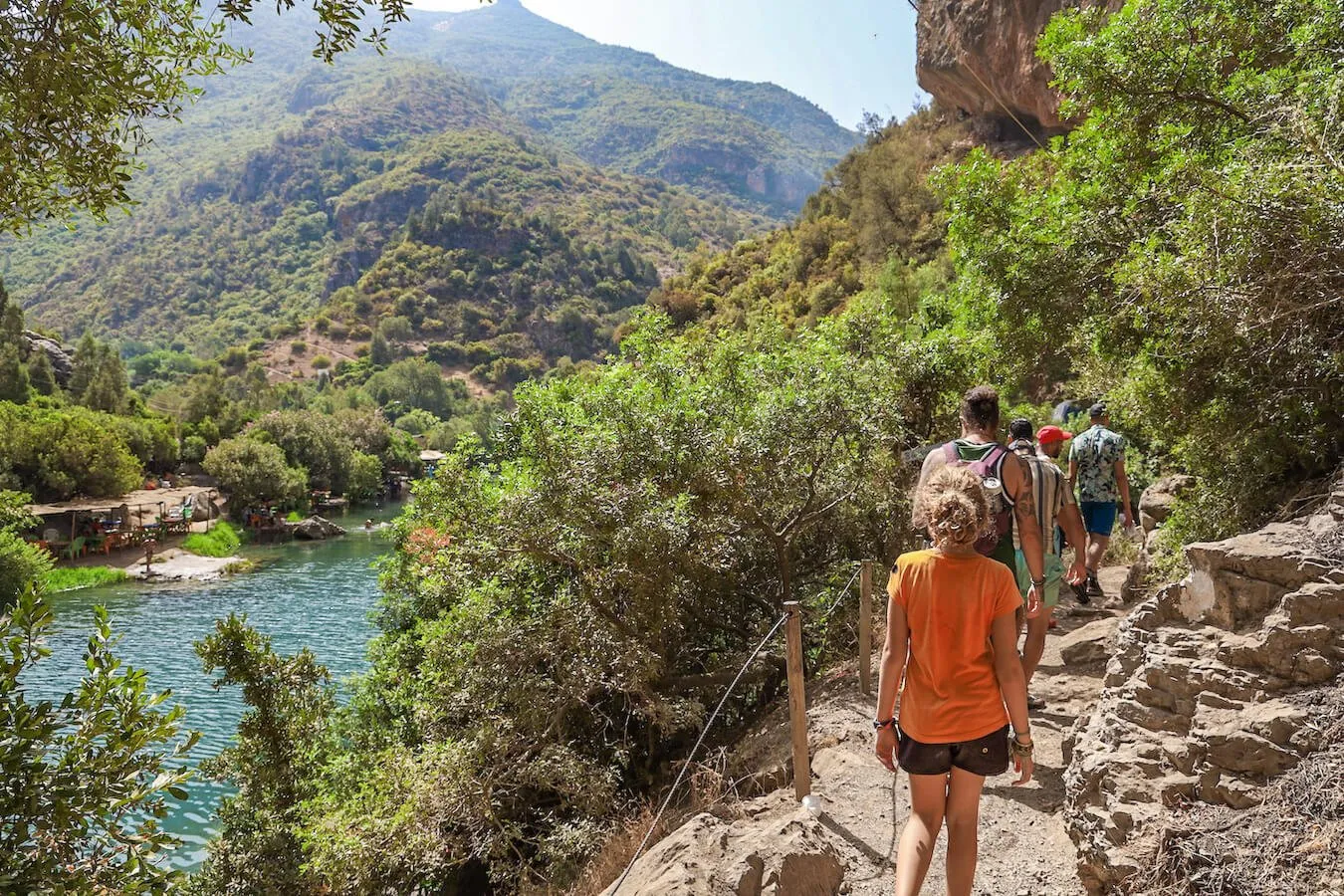 Group of hikers walking on a trail beside a river surrounded by green trees and mountains.